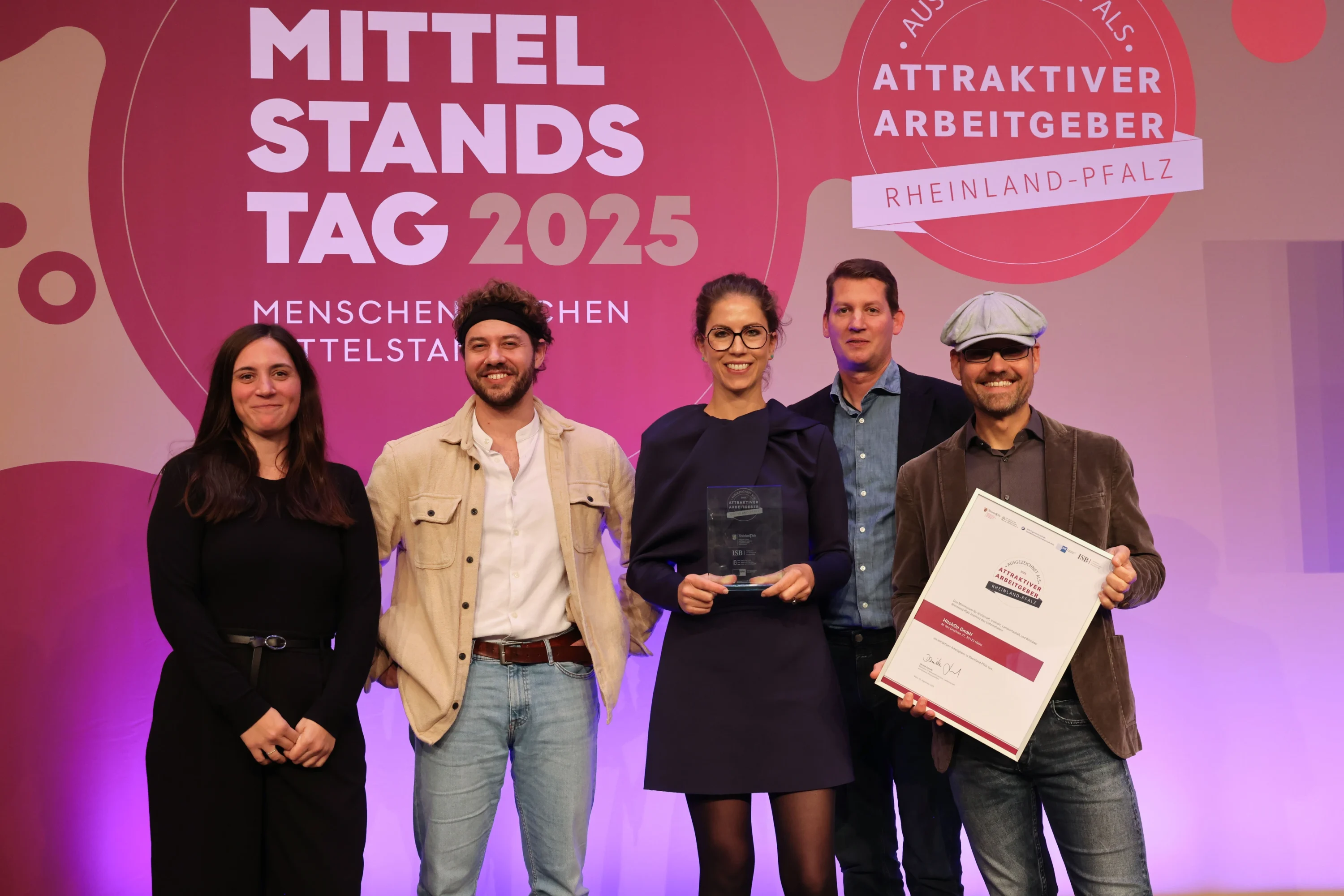 2 Frauen und 3 Männer stehen vor einer Plakatwand. Einer der Männer ganz rechts hält eine Urkunde.